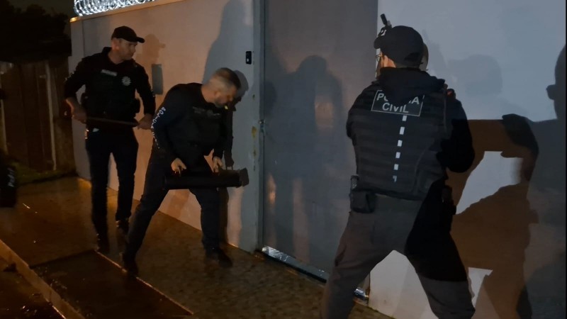 Três agentes fardados da Polícia Civil se preparam para arrombar uma porta de metal na operação Cifra Oculta