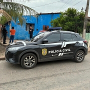 A imagem apresenta em primeiro plano um carro da PC estacionado em frente a uma casa azul, quatro pessoas conversam, sendo dois fardados