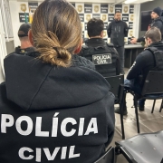 A foto mostra agentes da Polícia Civil de costas, com o nome da corporação em seus uniformes. Eles estão sentados em uma sala, disposta no formato auditório, e à frente, dois policiais falam voltados para os demais. Há um fundo de palco quadriculado com imagens alternadas do logotipo da Polícia Civil e o nome da instituição.