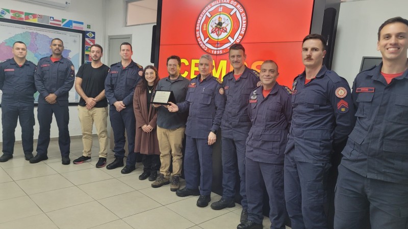 Há 11 pessoas na foto, oito delas com farda azul dos bombeiros, e outras três com roupas civis. Elas estão à frente de um painel onde está projetado o logotipo do Corpo de Bombeiros Militar do Rio Grande do Sul, com fundo vermelho.
