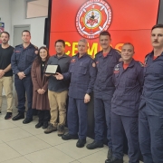 Há 11 pessoas na foto, oito delas com farda azul dos bombeiros, e outras três com roupas civis. Elas estão à frente de um painel onde está projetado o logotipo do Corpo de Bombeiros Militar do Rio Grande do Sul, com fundo vermelho.