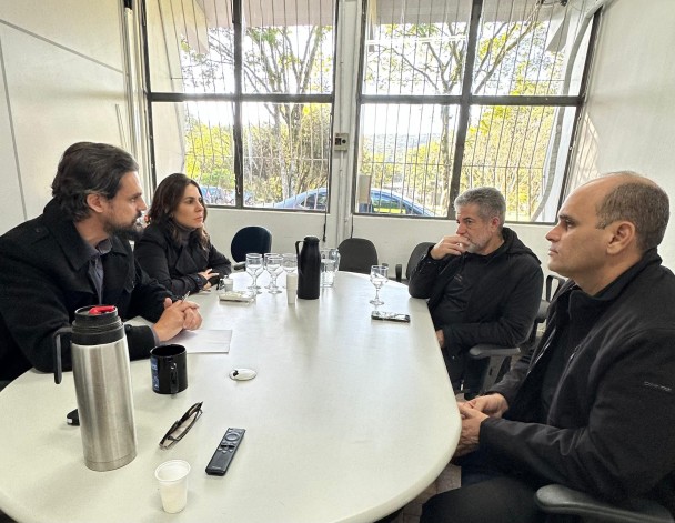 Quatro pessoas estão reunidas em torno de uma mesa, com garrafas térmicas e outros objetos, como celulares, óculos, taças e copos. 