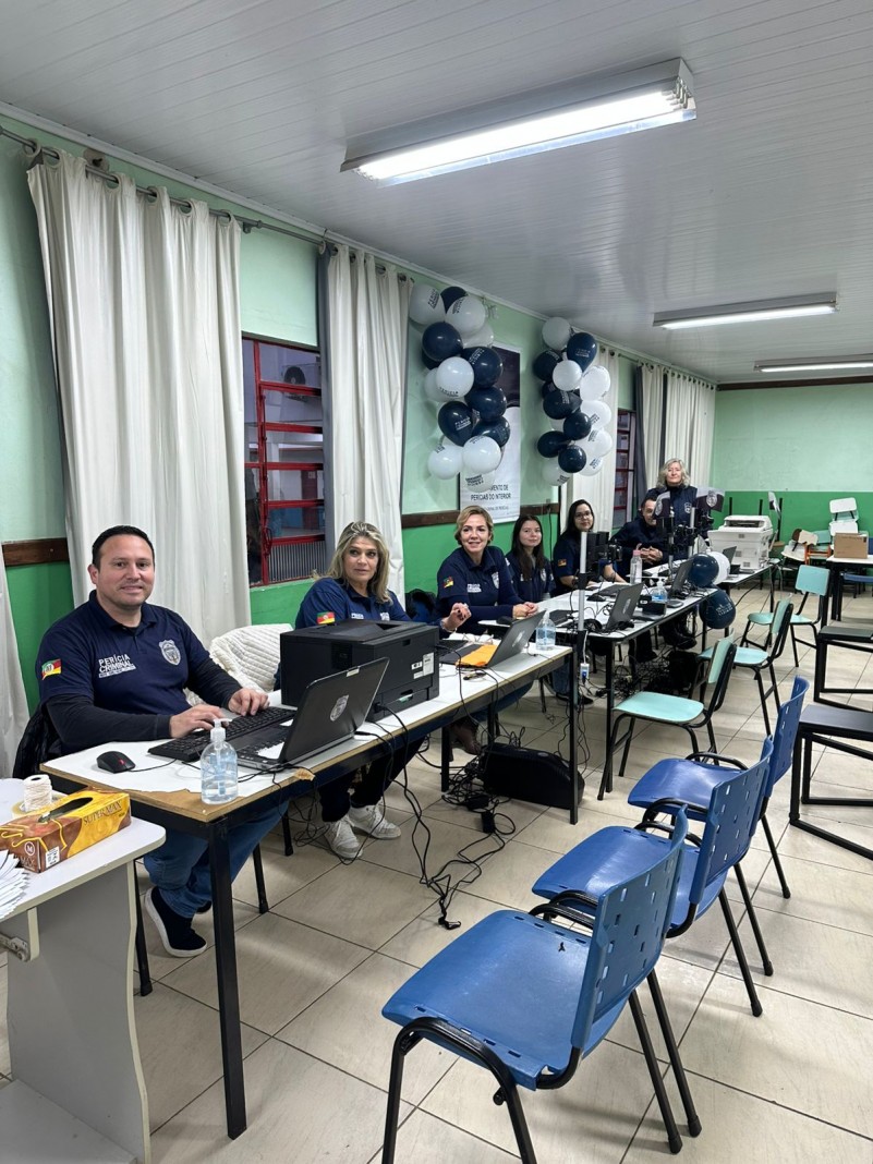 Sete agentes do IGP estão sentados lado a lado, cada um com uma mesa e um notebook à frente, em um ambiente parecido com uma sala de aula grande. À frente, cadeiras para atendimento.