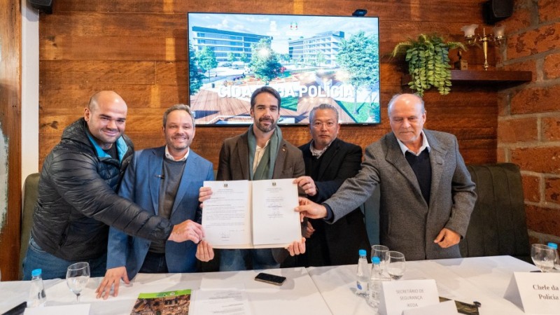 A imagem mostra, da esquerda para a direita, o presidente da Câmara de Vereadores de Passo Fundo, o prefeito de Passo Fundo, Pedro Almeida, o governador Eduardo Leite, o secretário adjunto Mário Ikeda e o chefe de Polícia, Heraldo Guerreiro. Todos seguram e mostram para as câmeras o termo de cooperação assinado