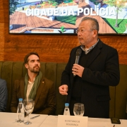 A imagem mostra o secretário adjunto Mário Ikeda em pé, segurando um microfone, enquanto discursa no evento. Ao lado, o governador Eduardo Leite está sentado e olha para o secretário adjunto 