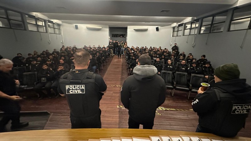 A imagem mostra três agentes da Polícia Civil de costas para a câmera e de frente para uma plateia composta por outros agentes. Os três policiais estão de pé, enquanto os demais agentes estão sentados. Ao fundo da sala, há algumas pessoas em pé