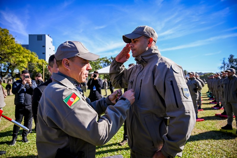 A imagem mostra o subcomandante-geral da Brigada Militar, coronel Douglas da Rosa Soares, condecorando um dos alunos do curso. O aluno está em pé, prestando continência, enquanto recebe a medalha
