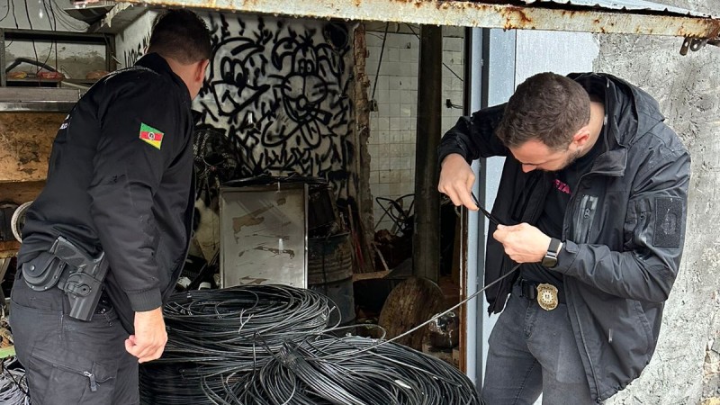 A imagem mostra dois agentes das for&ccedil;as de seguran&ccedil;a examinando fios apreendidos durante a opera&ccedil;&atilde;o. Os dois agentes est&atilde;o em p&eacute; e vestem jaquetas pretas. Os fios apreendidos est&atilde;o em cima de uma estrutura met&aacute;lica para que serve de transporte do material