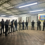 A imagem mostra agentes das forças de segurança reunidos num ambiente interno do Batalhão da Brigada Militar em São Leopoldo. Todos estão em pé, em posição de descanso, enquanto recebem instruções antes do início da operação
