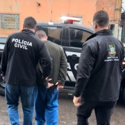 A imagem mostra um homem preso em flagrante sendo conduzido para uma viatura da Polícia Civil. Ele está acompanhado por dois agentes das forças de segurança. O agente à esquerda veste uma jaqueta da cor preta da Polícia Civil. Um pouco atrás do homem detido, está um agente vestindo uma jaqueta da cor preta da Secretaria da Segurança Pública do RS