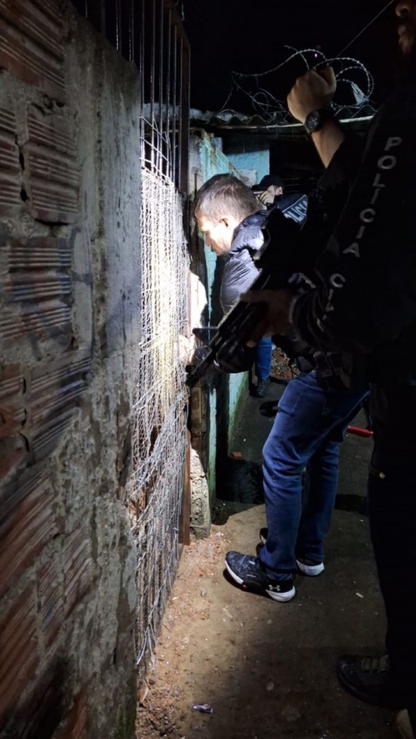 Agente entra em porta esteira na madrugada, iluminado por um facho de lanterna de outro agente, que vem logo atrás