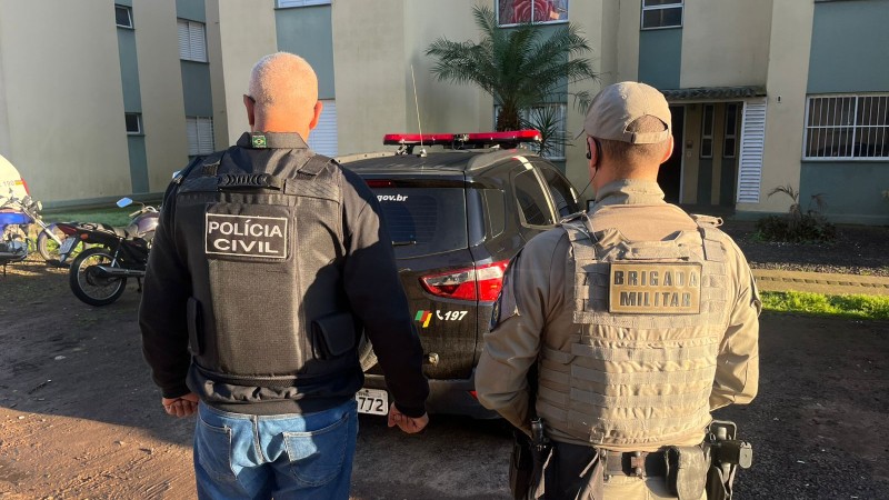 Imagem mostra dois policiais de costas e em frente a um pr&eacute;dio de apartamentos.