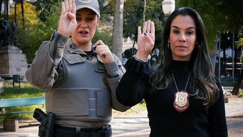 Duas mulheres fazem o sinal para identificar mulheres em situa&ccedil;&atilde;o de viol&ecirc;ncia. Elas est&atilde;o com a m&atilde;o direita erguida, com o polegar dobrado sobre a m&atilde;o e os quatro dedos para cima.