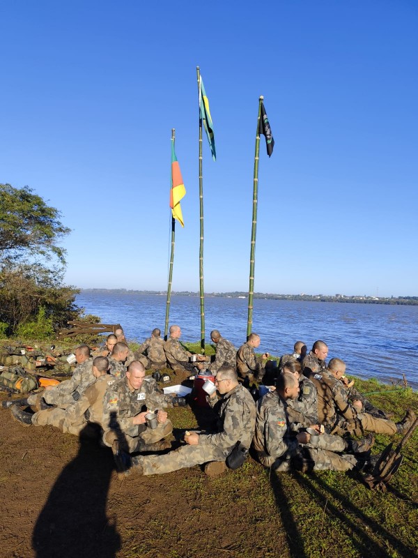 A imagem mostra os alunos do  Curso de Especialização em Operações Ribeirinhas sentados à margem de um rio durante um dos exercícios práticos do curso. É possível ver, hasteadas em mastros de bambu, bandeiras do Rio Grande do Sul, do Brasil e do 6º BPChq. Os alunos vestem militares fardas camufladas
