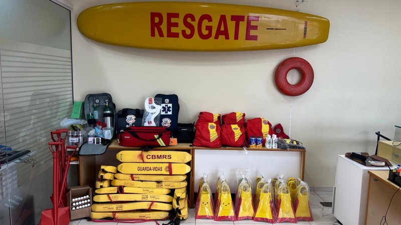 A imagem mostra uma sala com diversos equipamentos de salvamento utilizados por guarda-vidas. Na parede, h&aacute; uma prancha de resgate amarela com a palavra &ldquo;RESGATE&rdquo; em vermelho. Ao lado, est&aacute; pendido um flutuador circular vermelho. Sobre uma mesa, encontram-se mochilas vermelhas, bolsas de primeiros socorros, cilindros de oxig&ecirc;nio, colar cervical, protetor solar e outros itens m&eacute;dicos. No ch&atilde;o, empilhados, h&aacute; v&aacute;rios flutuadores amarelos com a inscri&ccedil;&atilde;o &ldquo;CBMRS &ndash; GUARDA-VIDAS&rdquo;, al&eacute;m de pares de nadadeiras tamb&eacute;m nas cores amarela e vermelha embaladas em pl&aacute;stico. &Agrave; esquerda, aparecem p&aacute;s e outros materiais de apoio.