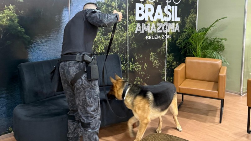 A imagem mostra um policial militar e um c&atilde;o realizando uma vistoria de seguran&ccedil;a preventiva em um ambiente fechado durante um evento. 