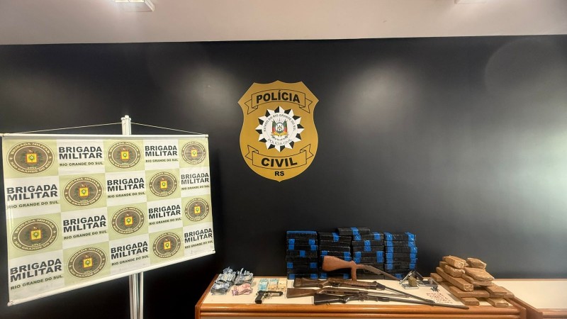 Esta &eacute; uma descri&ccedil;&atilde;o detalhada da imagem para fins de acessibilidade: Vis&atilde;o Geral A imagem mostra a exibi&ccedil;&atilde;o de materiais apreendidos em uma opera&ccedil;&atilde;o policial conjunta entre a Pol&iacute;cia Civil e a Brigada Militar. Os itens est&atilde;o organizados sobre uma mesa de madeira contra uma parede preta, em um ambiente interno iluminado por l&acirc;mpadas fluorescentes no teto. Elementos de Fundo e Identifica&ccedil;&atilde;o Parede Central: Uma parede pintada de preto fosco exibe, ao centro, um grande emblema dourado e preto da Pol&iacute;cia Civil do RS. Banner &agrave; Esquerda: Um banner vertical (roll-up) com fundo branco apresenta o bras&atilde;o da Brigada Militar do Rio Grande do Sul repetido em um padr&atilde;o de grade verde e dourado. Materiais Apreendidos (sobre a mesa) Da esquerda para a direita, os itens est&atilde;o dispostos da seguinte forma: Dinheiro: V&aacute;rios ma&ccedil;os de c&eacute;dulas de Real (notas de diversos valores) organizados em pequenos montes. Armamento: * Uma pistola preta de pequeno porte. Tr&ecirc;s armas longas (espingardas ou rifles) com acabamento em madeira e metal, dispostas horizontalmente. Entorpecentes: Duas grandes pilhas de tijolos retangulares envoltos em fita adesiva azul, totalizando dezenas de unidades. Uma pilha menor de tijolos retangulares envoltos em fita adesiva parda (cor de papel&atilde;o), localizada no canto direito da mesa. Contexto A imagem ilustra os resultados da opera&ccedil;&atilde;o "Ver&atilde;o Sem Drogas", focada no combate ao tr&aacute;fico de entorpecentes na regi&atilde;o do Litoral Norte do estado.