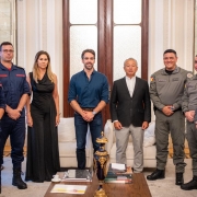 Esta &eacute; uma descri&ccedil;&atilde;o detalhada da imagem para fins de acessibilidade: Vis&atilde;o Geral A imagem registra um momento oficial em que oito pessoas &mdash; sete homens e uma mulher &mdash; posam para uma fotografia em um ambiente luxuoso e formal. O grupo est&aacute; organizado em uma linha reta, transmitindo uma atmosfera de coopera&ccedil;&atilde;o institucional. Pessoas e Vestimentas (da esquerda para a direita) Bombeiros Militares: Os dois primeiros homens &agrave; esquerda vestem uniformes operacionais azul-marinho com detalhes e identifica&ccedil;&otilde;es em vermelho, t&iacute;picos do Corpo de Bombeiros. Civil: Uma mulher com cabelos loiros longos veste um vestido preto longo e elegante. Governador: No centro da imagem, Eduardo Leite veste uma camisa social azul-marinho com as mangas levemente dobradas e cal&ccedil;a jeans escura. Civil/Autoridade: &Agrave; direita do governador, um homem de cabelos grisalhos veste um blazer preto, camisa branca e cal&ccedil;a cinza-clara. Brigada Militar: Os dois homens seguintes vestem fardas cinza-oliva da Brigada Militar, com diversos distintivos e ins&iacute;gnias. T&aacute;tico: Na extremidade direita, um homem veste um uniforme t&aacute;tico camuflado em tons de cinza e preto, equipado com colete operacional. Cen&aacute;rio e Composi&ccedil;&atilde;o O Ambiente: O encontro ocorre em uma sala de recep&ccedil;&atilde;o cl&aacute;ssica, possivelmente no Pal&aacute;cio Piratini. O fundo apresenta uma grande porta dupla de madeira com pain&eacute;is de vidro fosco, paredes brancas decoradas com boiseries (molduras de gesso) e espelhos laterais. Mobili&aacute;rio: Nas laterais, veem-se partes de sof&aacute;s brancos. &Agrave; frente do grupo, h&aacute; uma mesa de centro de madeira escura contendo livros, uma pequena x&iacute;cara de caf&eacute; sobre um pires branco e um vaso decorativo alongado em azul-marinho com detalhes dourados. Ilumina&ccedil;&atilde;o: A luz &eacute; suave e bem distribu&iacute;da, real&ccedil;ando os detalhes dos uniformes e do ambiente.