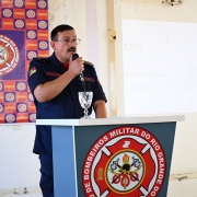 O corregedor geral dos bombeiros militares está em pé atrás de um púlpito com o brasão do Corpo de Bombeiros Militar do Rio Grande do Sul. Ele segura um microfone próximo à boca, como se estivesse discursando. Sobre o púlpito, há uma taça decorativa. Ao fundo, vê-se um painel com repetição do emblema da corporação e, ao lado, uma projeção clara na parede, indicando o uso de recursos audiovisuais durante a fala.