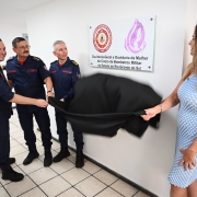 Em um corredor interno de paredes claras, três bombeiros militares e uma mulher participam do descerramento de uma placa fixada na parede. Todos seguram um tecido escuro que está sendo retirado da placa. A placa contém textos e símbolos institucionais, incluindo o brasão do Corpo de Bombeiros Militar e referências à Ouvidoria-Geral e à Ouvidoria da Mulher. Outras pessoas acompanham o momento, e uma delas registra a cena com um celular.