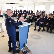 Em uma sala ampla e bem iluminada, um integrante do Corpo de Bombeiros Militar está em pé atrás de um púlpito azul, falando ao público e gesticulando com as mãos. Ele usa uniforme azul-escuro. À sua frente, várias pessoas estão sentadas em fileiras de cadeiras, muitas delas também vestindo uniformes semelhantes, voltadas para o orador e ouvindo atentamente. Ao fundo, há janelas grandes que deixam entrar luz natural, além de mesas, equipamentos e outros participantes em pé acompanhando a apresentação.