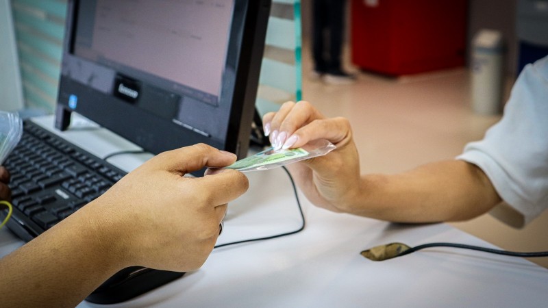 Em primeiro plano, uma pessoa est&aacute; entregando um cart&atilde;o plastificado, um documento de identidade, para outra pessoa. O cart&atilde;o &eacute; segurado com a m&atilde;o estendida, sugerindo um momento de verifica&ccedil;&atilde;o ou registro. Ao fundo, h&aacute; um computador de mesa com teclado e monitor, refor&ccedil;ando o contexto administrativo ou oficial da intera&ccedil;&atilde;o. O cen&aacute;rio transmite a ideia de um processo formal de atendimento ou confer&ecirc;ncia de documentos.