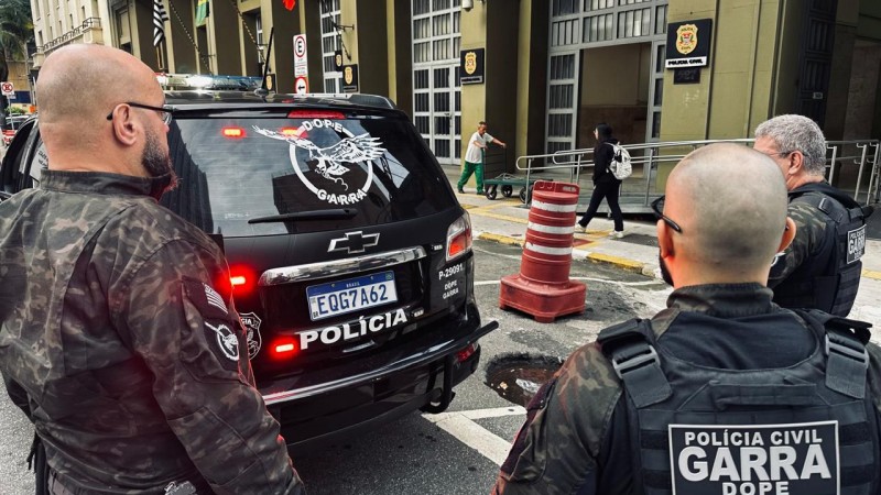 Tr&ecirc;s policiais da unidade GARRA, da Pol&iacute;cia Civil, aparecem em frente a uma viatura preta com emblemas da corpora&ccedil;&atilde;o e da unidade especial. Eles vestem uniformes camuflados e equipamentos t&aacute;ticos. Ao fundo, h&aacute; um pr&eacute;dio oficial da Pol&iacute;cia Civil, bandeiras do Brasil e de S&atilde;o Paulo, al&eacute;m de pessoas circulando, indicando um ambiente urbano.