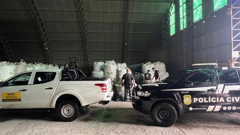 Interior de um galp&atilde;o. No fundo, grandes sacos brancos empilhados. Em primeiro plano, dois ve&iacute;culos: uma caminhonete branca com faixa amarela escrita &ldquo;MAPA &ndash; Minist&eacute;rio da Agricultura, Pecu&aacute;ria e Abastecimento &ndash; Uso exclusivo em servi&ccedil;o&rdquo; e uma viatura preta da Pol&iacute;cia Civil com logotipo e endere&ccedil;o de redes sociais. Algumas pessoas em uniforme est&atilde;o pr&oacute;ximas aos sacos, realizando inspe&ccedil;&atilde;o.