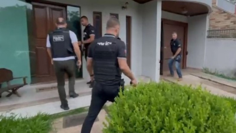 Exterior de uma casa moderna. H&aacute; um caminho de piso claro e um arbusto verde ao lado da entrada. V&aacute;rios policiais, com coletes pretos escritos &ldquo;POL&Iacute;CIA&rdquo;, aproximam-se da porta. Um deles usa uma ferramenta para abrir a porta, enquanto outros observam e se preparam para entrar. A cena mostra uma opera&ccedil;&atilde;o policial em andamento. 