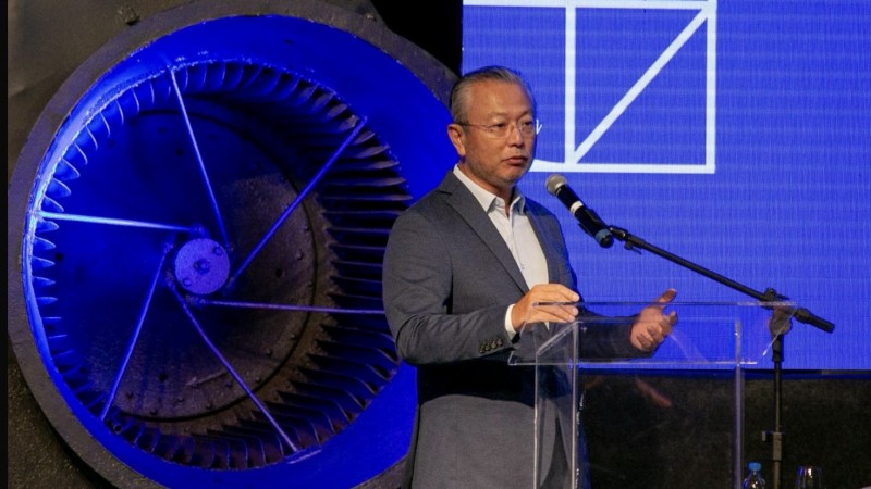 A imagem mostra um homem de terno falando em um p&uacute;lpito com microfone, em um ambiente de evento formal. Atr&aacute;s dele h&aacute; uma grande estrutura met&aacute;lica iluminada em azul, lembrando uma turbina, que d&aacute; um aspecto industrial ao cen&aacute;rio. &Agrave; direita, outro homem de terno claro aparece parcialmente de costas. No fundo, uma tela azul com desenhos geom&eacute;tricos brancos comp&otilde;e a apresenta&ccedil;&atilde;o. Sobre a mesa pr&oacute;xima ao p&uacute;lpito h&aacute; uma garrafa de &aacute;gua e um copo.