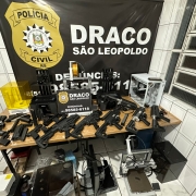 A imagem mostra uma opera&ccedil;&atilde;o da Pol&iacute;cia Civil do Rio Grande do Sul, realizada pela unidade DRACO de S&atilde;o Leopoldo. Em destaque, h&aacute; um banner institucional com o nome da corpora&ccedil;&atilde;o e um n&uacute;mero de contato para den&uacute;ncias. &Agrave; frente do banner, sobre uma mesa, est&atilde;o expostos diversos itens apreendidos: armas de fogo, componentes de armas e v&aacute;rias impressoras 3D, sugerindo que a tecnologia estava sendo utilizada para fabricar ou modificar armamentos. O cen&aacute;rio transmite claramente o car&aacute;ter de uma a&ccedil;&atilde;o policial voltada ao combate &agrave; produ&ccedil;&atilde;o e distribui&ccedil;&atilde;o ilegal de armas.