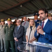 A imagem registra um evento oficial em que o Governador do Rio Grande do Sul, Eduardo Leite, discursa para uma plateia de autoridades civis e militares. Composi&ccedil;&atilde;o da Imagem Primeiro Plano: &Agrave; direita, Eduardo Leite aparece em destaque, falando ao microfone em um p&uacute;lpito de acr&iacute;lico transparente. Ele tem cabelos e barba aparados, veste um palet&oacute; azul marinho, camisa branca e gravata bord&ocirc;. Ele segura o microfone com as duas m&atilde;os e olha para o lado oposto &agrave; c&acirc;mera. Segundo Plano: Logo atr&aacute;s do governador, diversas autoridades acompanham o discurso. Est&atilde;o presentes homens e mulheres em trajes formais (ternos e vestidos) e v&aacute;rios oficiais militares de alta patente, vestindo fardas em tons de verde-oliva e azul, adornadas com ins&iacute;gnias e medalhas. Ambiente: A cerim&ocirc;nia ocorre em um local aberto, mas sob uma cobertura de estrutura met&aacute;lica aparente. A ilumina&ccedil;&atilde;o &eacute; natural, sugerindo um evento realizado durante o dia em um p&aacute;tio ou pavilh&atilde;o. Clima e Tom A cena transmite uma atmosfera de solenidade e formalidade, t&iacute;pica de cerim&ocirc;nias de posse e an&uacute;ncios governamentais. As express&otilde;es dos presentes s&atilde;o s&eacute;rias e atentas.