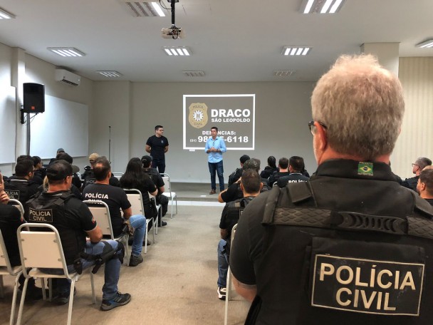Em São Leopoldo, agentes uniformizados da Polícia Civil assistem a uma apresentação em sala equipada com projetor e tela. Dois palestrantes estão à frente, e o conteúdo exibido reforça a identidade da DRACO e divulga um número de contato para denúncias: 51 98585 6118.