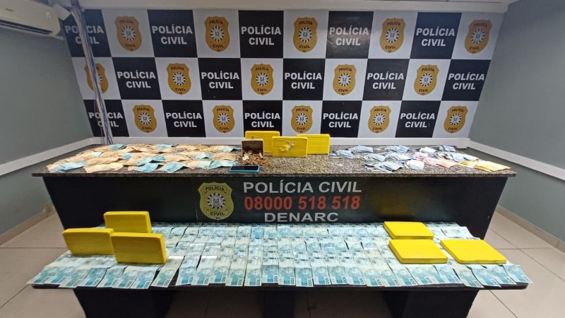 Uma mesa com diversos itens confiscados: pacotes amarelos retangulares que aparentam conter subst&acirc;ncias il&iacute;citas, dinheiro em esp&eacute;cie, moedas em uma caixa e um celular, tudo exposto diante do painel da Pol&iacute;cia Civil e da divis&atilde;o DENARC, especializada em narc&oacute;ticos.