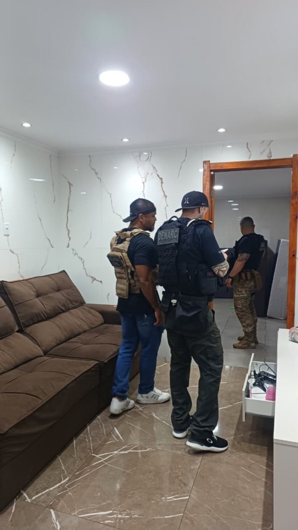 Três agentes do DENARC, equipados com coletes táticos, realizam uma busca dentro de uma residência moderna, com sofá, móveis e eletrônicos visíveis, reforçando o caráter de uma operação policial em ambiente doméstico.