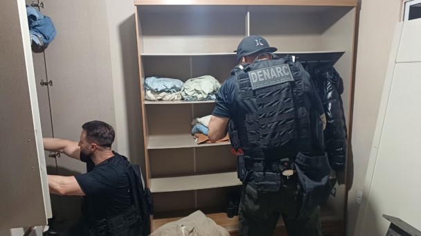 Dois policiais revistam detalhadamente um guarda-roupa, um deles ajoelhado examinando a parte inferior e outro em pé verificando roupas e prateleiras, evidenciando o processo minucioso de investigação em busca de drogas ou objetos relacionados ao tráfico.