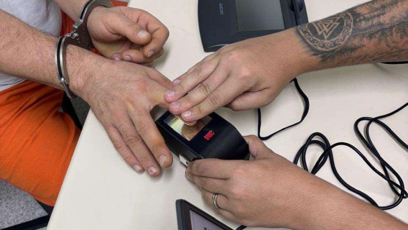 Foto mostra o detalhe da m&atilde;o de um detento colocando o dedo no leitor digital com ajuda do servidor. Ele est&aacute; algemado e aparece parte da cal&ccedil;a laranja do uniforme.