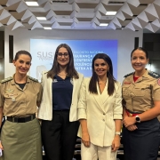 Quatro mulheres est&atilde;o em p&eacute;, lado a lado, em uma sala de confer&ecirc;ncia ou semin&aacute;rio. Duas delas vestem uniformes de for&ccedil;as de seguran&ccedil;a (militares ou policiais), enquanto as outras duas usam roupas profissionais civis. Ao fundo, h&aacute; uma tela de proje&ccedil;&atilde;o exibindo um slide com o texto em portugu&ecirc;s:
"SUS Mulheres &ndash; 1&ordm; Encontro Nacional &ndash; Seguran&ccedil;a P&uacute;blica e o Enfrentamento da Viol&ecirc;ncia Contra a Mulher". O ambiente transmite formalidade e colabora&ccedil;&atilde;o entre representantes da seguran&ccedil;a p&uacute;blica e da sociedade civil.