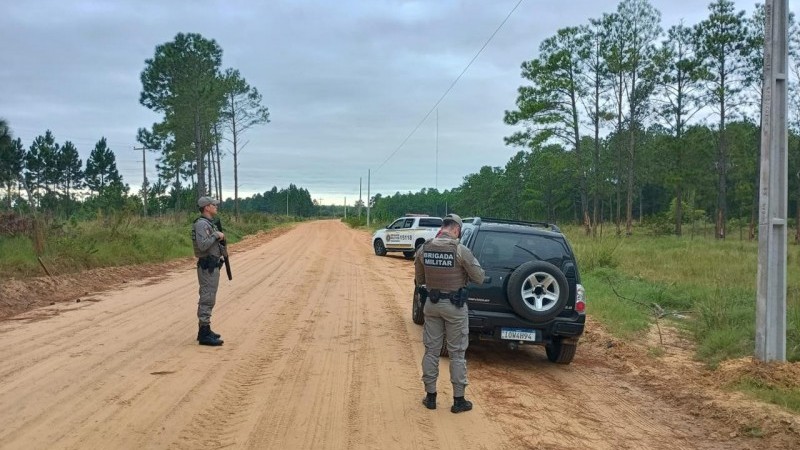 A imagem mostra dois policiais militares realizando patrulhamento em uma &aacute;rea rural. &Eacute; poss&iacute;vel ver um ve&iacute;culo de passeio e uma viatura da Brigada Militar em uma estrada de terra