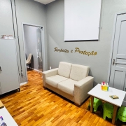 Um ambiente acolhedor e organizado, com paredes cinza claro e piso de madeira. H&aacute; um sof&aacute; bege de dois lugares encostado na parede, acima do qual est&aacute; escrito em letras douradas &ldquo;Respeito e Prote&ccedil;&atilde;o&rdquo;. Ao lado, uma mesa branca pequena com dois banquinhos verdes em forma de elefante, sobre a qual est&atilde;o brinquedos e um livro. Um vaso grande com planta verde est&aacute; pr&oacute;ximo &agrave; porta branca, e do outro lado h&aacute; prateleiras com livros, brinquedos e uma boneca. O espa&ccedil;o transmite sensa&ccedil;&atilde;o de cuidado e seguran&ccedil;a, possivelmente pensado para crian&ccedil;as ou fam&iacute;lias.