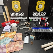 Uma mesa repleta de itens apreendidos pela Pol&iacute;cia Civil RS, especificamente da unidade DRACO Passo Fundo. Entre os objetos expostos est&atilde;o dois rev&oacute;lveres, muni&ccedil;&otilde;es, diversos celulares, balan&ccedil;as digitais, rolos de pl&aacute;stico filme, pacotes suspeitos, pen drives e uma quantia significativa de dinheiro em notas brasileiras. Ao fundo, dois pain&eacute;is pretos exibem o nome da institui&ccedil;&atilde;o e contatos, incluindo um n&uacute;mero de WhatsApp para den&uacute;ncias, refor&ccedil;ando o car&aacute;ter oficial da opera&ccedil;&atilde;o e a divulga&ccedil;&atilde;o p&uacute;blica dos resultados da apreens&atilde;o.
