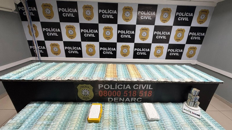 A imagem mostra, sobre uma mesa, grande quantidade de notas de 100 reais empilhadas e organizadas em ma&ccedil;os. Tamb&eacute;m aparecem pacotes retangulares embrulhados em material branco, al&eacute;m de uma balan&ccedil;a digital com mais dinheiro em cima. No fundo, a parede tem o padr&atilde;o quadriculado preto e branco com o logotipo e o texto &ldquo;POL&Iacute;CIA CIVIL&rdquo;, acompanhado do bras&atilde;o dourado da corpora&ccedil;&atilde;o. Na frente da mesa, h&aacute; uma faixa com os dizeres &ldquo;POL&Iacute;CIA CIVIL 08000 518 518 DENARC&rdquo;, indicando que se trata de uma opera&ccedil;&atilde;o do departamento de narc&oacute;ticos. Essa composi&ccedil;&atilde;o sugere que a foto foi tirada em uma delegacia, como registro oficial de uma apreens&atilde;o de dinheiro e poss&iacute;veis materiais relacionados a investiga&ccedil;&atilde;o.