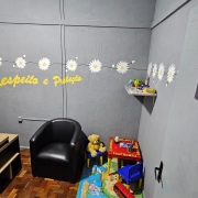 Sala de atendimento identificada como &ldquo;Sala das Margaridas&rdquo;, com decora&ccedil;&atilde;o acolhedora e tem&aacute;tica infantil. O espa&ccedil;o inclui poltrona, mesa de trabalho, brinquedos, tapete colorido e prateleira com objetos. Na parede, h&aacute; o texto &ldquo;Respeito e Prote&ccedil;&atilde;o&rdquo; em amarelo, al&eacute;m de flores decorativas.