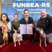 Grupo de quatro pessoas posando sobre um tapete vermelho em frente a um painel com o texto &ldquo;FUNBEA-RS&rdquo;, exibindo um documento aberto para a c&acirc;mera; tr&ecirc;s c&atilde;es aparecem em primeiro plano ao lado dos participantes.