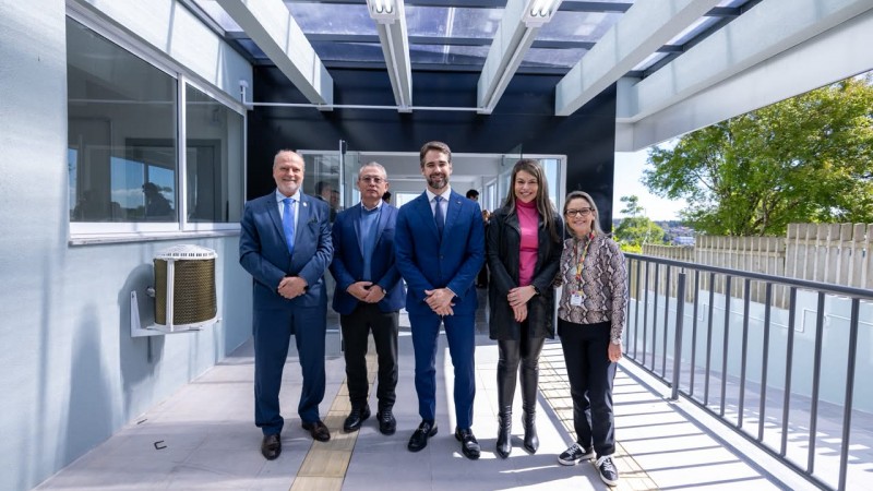 Grupo de pessoas posando em uma passarela coberta de um edif&iacute;cio moderno, com paredes em tons claros, cobertura transl&uacute;cida e guarda-corpo met&aacute;lico; ao fundo, h&aacute; portas envidra&ccedil;adas e vista parcial da &aacute;rea externa.