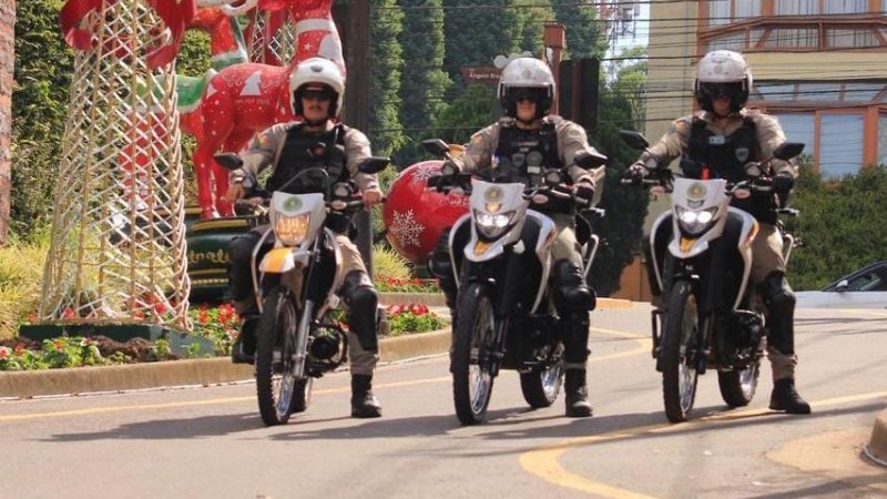 A imagem mostra tr&ecirc;s policias fazendo rondas com as motocicletas da Brigada Militar em uma rua da cidade de Gramado. &Eacute; poss&iacute;vel ver decora&ccedil;&otilde;es de Natal ao fundo